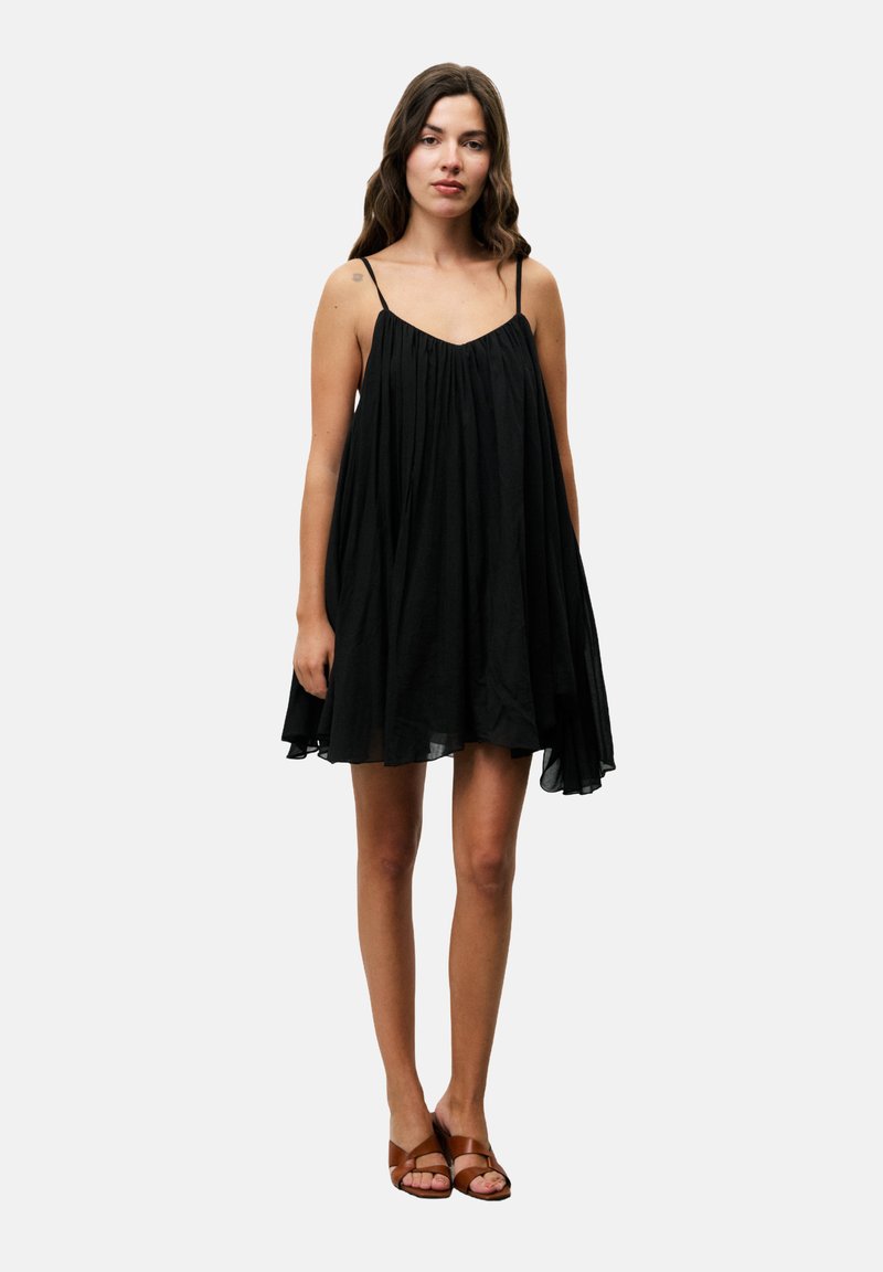 Zwarte mouwloze jurk met een vloeiende silhouette, gathering bij de buste en een zachte textuur. Draagt met bruine sandalen met banden.