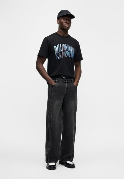 Μαύρο μπλουζάκι με μπλε γραφικό κείμενο, φαρδιά ξεθωριασμένα μαύρα τζιν και μαύρα παπούτσια. Το σύνολο έχει χαλαρή σιλουέτα και διακριτικές λεπτομέρειες.