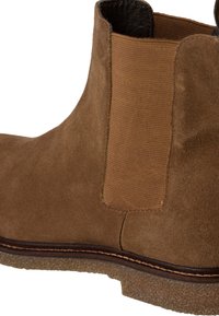Bota de ante marrón hasta el tobillo con panel lateral elástico, punta redondeada, suela de goma y detalle de costura en contraste. Textura suave, diseño resistente.