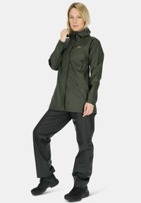 Vrouw met een donkergroene regenjas en zwarte regenbroek, met zwarte wandelschoenen, staand met een gebogen been en hand die de capuchon aanpast.