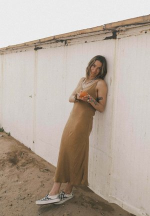 Beżowa, bezrękawnikowa suknia maxi o zwiewnym kroju; noszona z czarno-białymi wsuwanymi butami w kratę; uzupełniona warstwowymi naszyjnikami.