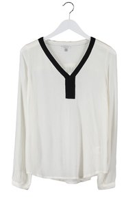 Blouse blanche à manches longues en tissu texturé, avec un col en V et une bordure noire autour du col. Suspendue sur un cintre.