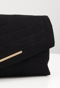 Černá semišová kabelka přes rameno s diagonálním prošíváním, zlatým kovovým detailem a sklápěcí klopou. Měkká textura a minimalistický design.