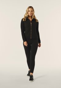 Gilet zipato nero con texture a trecce, abbinato a leggings neri e scarpe da ginnastica nere. Materiale morbido e design aderente.