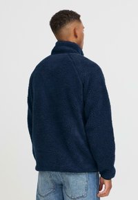 Marineblauer Fleece-Pullover mit hohem Kragen, strukturiertem, weichem Stoff und überschnittenen Schultern, kombiniert mit hellblauen Jeans.
