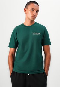 Grünes Baumwoll-T-Shirt mit kurzen Ärmeln und rundem Halsausschnitt. Merkmale ein weißes Grafiklogo auf der linken Brustseite. Glatte Textur.