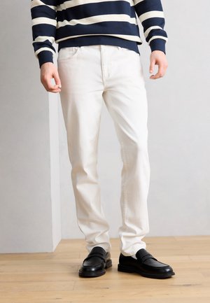 Άτομο που φοράει λευκά τζιν, μαύρα μοκασίνια και πουλόβερ με μπλε ναυτικό και λευκές ρίγες, στέκεται σε ελαφρύ ξύλινο πάτωμα.