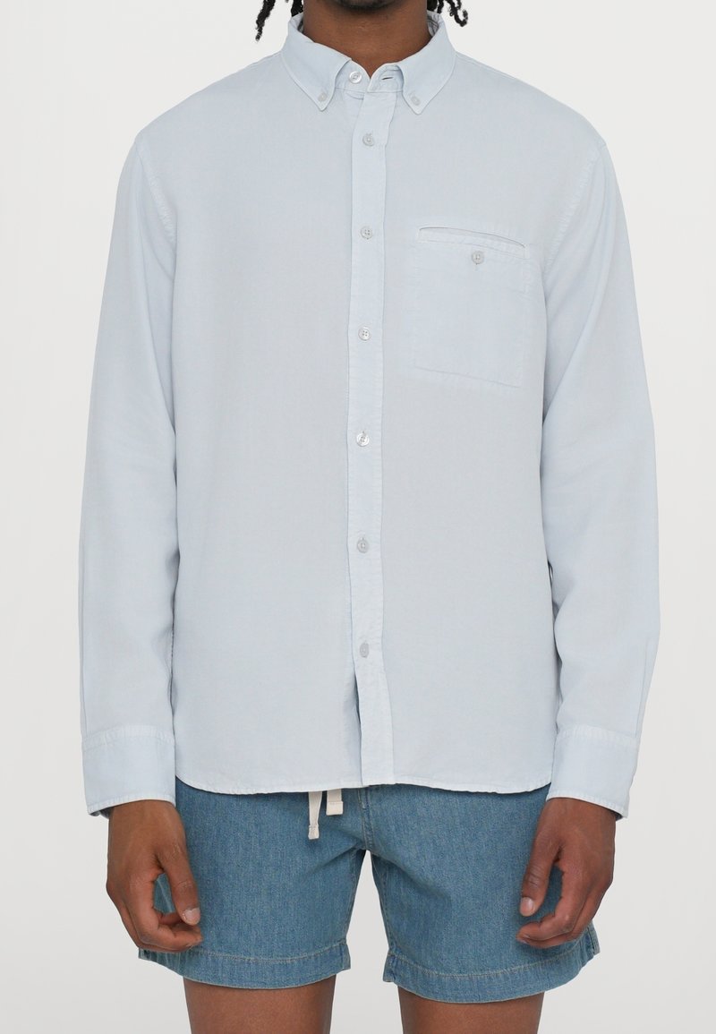 Světlomodrá košile se zapínáním na knoflíky, vyrobená z měkké látky, s náprsní kapsou, dlouhými rukávy a límečkem zapínaným na knoflíky. Pod ní jsou krátké džínové šortky.