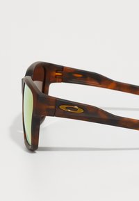 Occhiali da sole tartarugati con lenti verdi, dotati di un accento dorato sul lato e un design della montatura robusto e angolare.