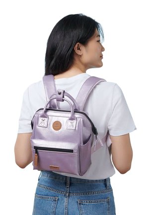 Frau trägt einen kleinen metallisch-lavendelfarbenen Rucksack mit zwei Griffen und einer Vorderreißverschlusstasche, kombiniert mit einem weißen Hemd und blauen Jeans.