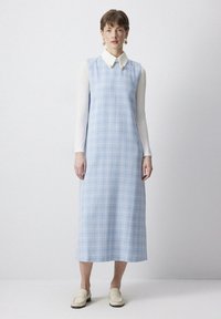 Rochie fără mâneci de culoare albastru deschis cu un model în carouri, având o cămașă albă cu guler dedesubt și mocasini simpli bej.