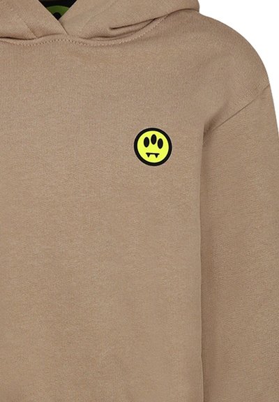 Felpa beige realizzata in materiale morbido, con una toppa rotonda nera raffigurante un sorriso e denti aguzzi in giallo brillante. Finitura con tessuto testurizzato.