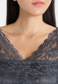 Top en dentelle grise avec des motifs floraux intricats, un col festonné et une texture douce, mettant en valeur les détails de la dentelle.
