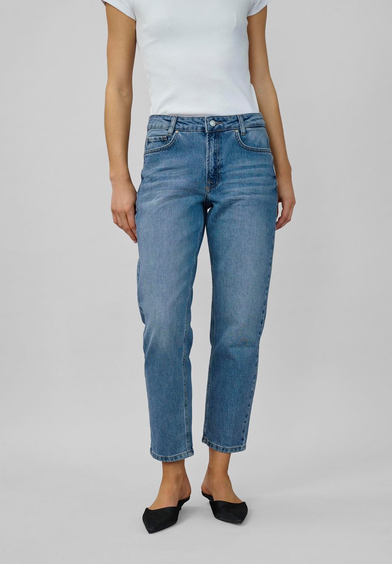 Femme portant un jean taille haute bleu, un haut blanc à manches courtes et des chaussures noires à bout pointu sans lacets, se tenant sur un fond gris uni.