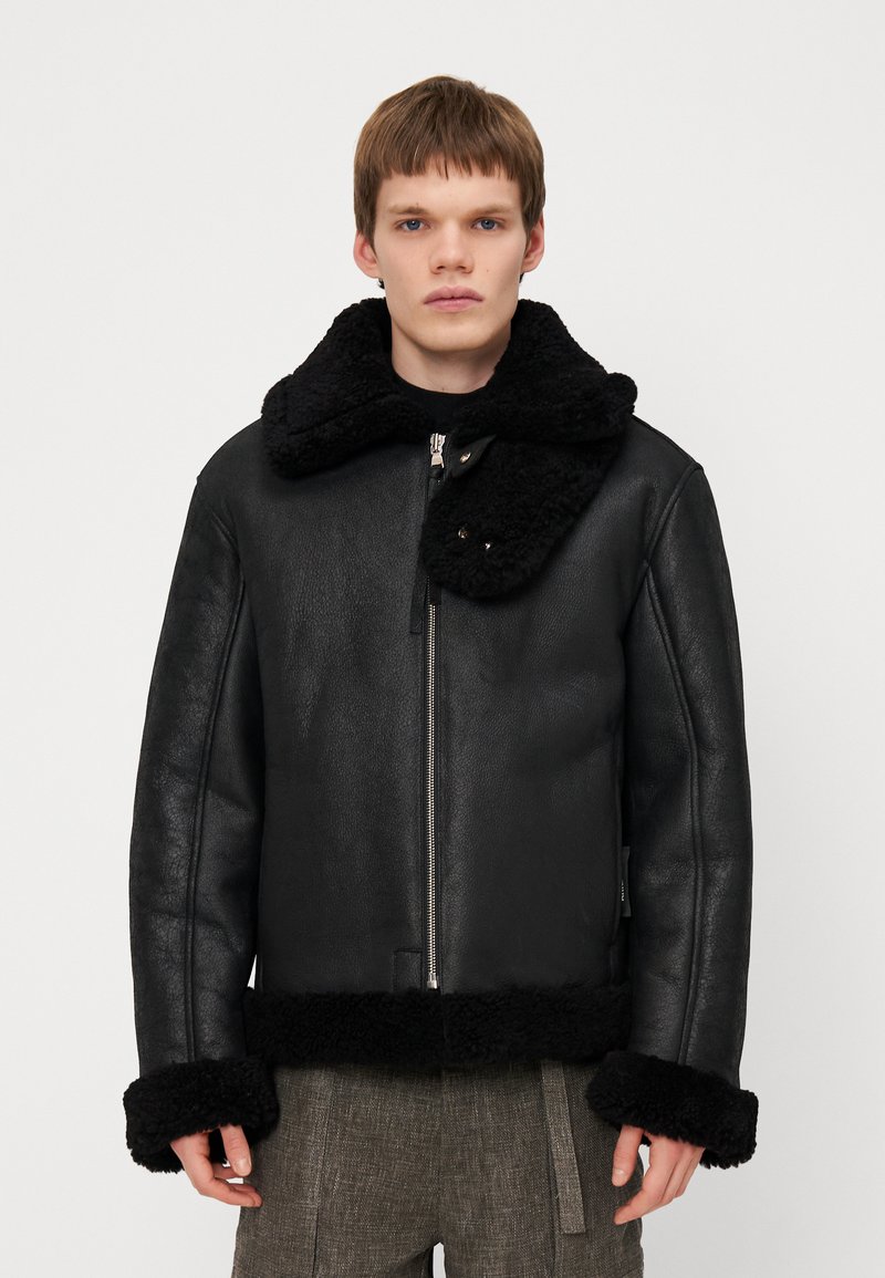 Chaqueta de cuero negra con un collar y puños de terciopelo negro. Presenta un cierre de cremallera en la parte delantera y un diseño de corte recto. El material parece suave con un acabado mate.