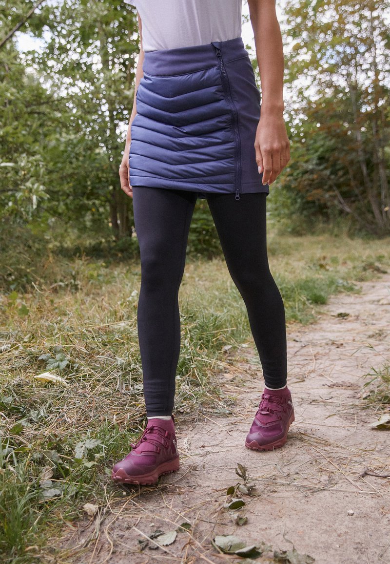 Padded, ciemnoniebieska spódnica z ukośnym pikowaniem i bocznym zamkiem, połączona z czarnymi legginsami i bordowymi butami turystycznymi. Naturalne tło.