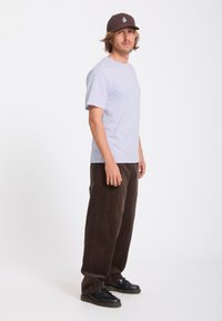 Homme portant un t-shirt gris clair, un pantalon ample marron foncé, des chaussures noires et une casquette marron foncé, debout sur un fond blanc.