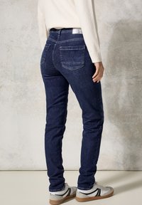 Tummansiniset farkut, joissa on slim fit -leikkaus, takaosassa taskut ja korkea vyötärö. Yhdistetty valkoisiin tennareihin, joissa on mustia ja harmaita yksityiskohtia.