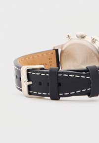 Μαύρο δερμάτινο λουράκι ρολογιού με λευκή ραφή και ασημένια αγκράφα, με μερική εμφάνιση της πίσω πλευράς μιας ασημένιας θήκης ρολογιού.