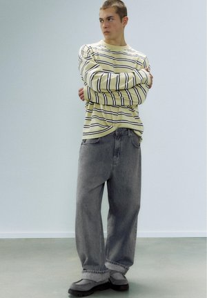 Junger Mann mit kurzen Haaren steht mit verschränkten Armen und trägt ein gelb gestreiftes Langarmshirt, lockere graue Jeans und graue Schuhe auf einem schlichten Boden.