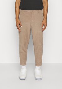 Uomo che indossa pantaloni in corduroy beige, sneaker bianche e calze bianche, in piedi davanti a uno sfondo bianco uniforme.