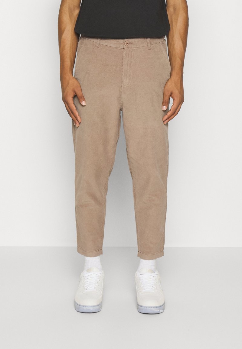 Uomo che indossa pantaloni in corduroy beige, sneaker bianche e calze bianche, in piedi davanti a uno sfondo bianco uniforme.