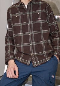 Chemise à carreaux marron à boutons avec des rayures vertes et blanches, dotée de deux poches poitrine et de boutons métalliques. Portée avec un pantalon cargo bleu.