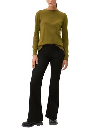 Vrouw in een olijfgroene lange mouwen trui, zwarte hoge taille wijde broek en zwarte chunky-soled laarzen, staande tegen een witte achtergrond.