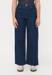 Jean large bleu marine à taille haute, doté d'une fermeture éclair et de boutons en métal, de poches latérales et d'ourlets nets. Porté avec des chaussures argentées.