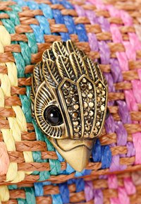 Spilla in bronzo a forma di uccello, con dettagli intricati delle piume, occhi neri e accenti scintillanti, posto su uno sfondo colorato di tessuto intrecciato.