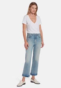 T-shirt bianco a V, jeans cropped azzurri chiari con strappi e orlo sfilacciato, e scarpe slip-on bianche con suola nera.