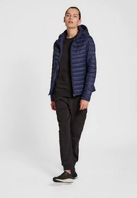 Granatowa pikowana kurtka z kapturem, zapinana na zamek, wykonana z lekkiego materiału o prostym kroju, zestawiona z czarnymi spodniami i butami.