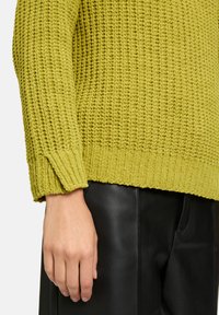 Kuscheliger Strickpullover in einem lebhaften Olivgrün, mit strukturiertem Rippenmuster und lockerer Passform. Kombiniert mit eleganten schwarzen Lederhosen.