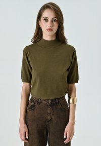 Maglietta a maniche corte in maglia verde oliva con collo alto, abbinata a pantaloni marroni a vita alta con texture e un bracciale in oro.