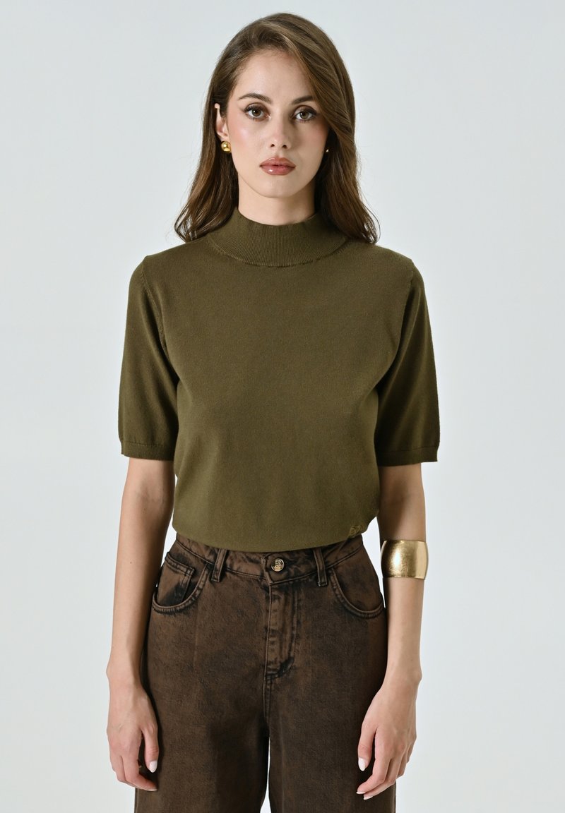 Maglietta a maniche corte in maglia verde oliva con collo alto, abbinata a pantaloni marroni a vita alta con texture e un bracciale in oro.