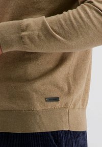 Brauner gestrickter Pullover mit gerippten Bündchen und Saum. Metallisches Logo-Tag an der Seite. Weicher Stoff, figurbetonter Schnitt und runder Halsausschnitt.