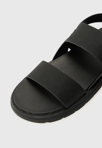 Svart sandal med tre brede stropper, flat såle og en jevn, matt finish. Sålen har en lett tekstur for bedre grep.