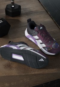Ζευγάρι παπουτσιών Adidas με μοτίβα μωβ, μαύρου και λευκού που ξεκουράζονται σε ξύλινο πάτωμα κοντά σε δύο μαύρα αλτήρες.