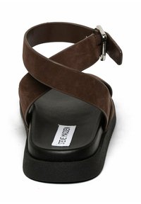 Sandales en faux daim marron avec des lanières entrecroisées, une semelle texturée noire et un accent de boucle en argent. L'étiquette indique "Steve Madden."
