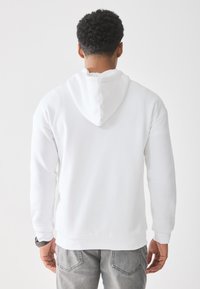 Een man met krullend haar, die met zijn rug naar de camera staat, draagt een eenvoudige witte hoodie en een grijze spijkerbroek, tegen een witte achtergrond.