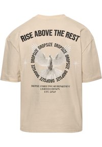 Beige bomulds T-shirt med korte ærmer. Ryggen har et grafisk motiv af en hvid due, omgivet af fed tekst og cirkulære designelementer.
