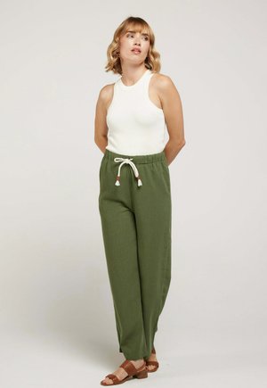 Femme debout, mains derrière le dos, portant un haut blanc sans manches, un pantalon olive à cordon et des sandales marron sur un fond beige.