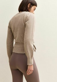 Beige gestrickte Strickjacke mit langen Ärmeln, Taillengürtel und geripptem Saum, kombiniert mit braunen, enganliegenden, gerippten Leggings. Strukturierte Stoffdetails sind sichtbar.