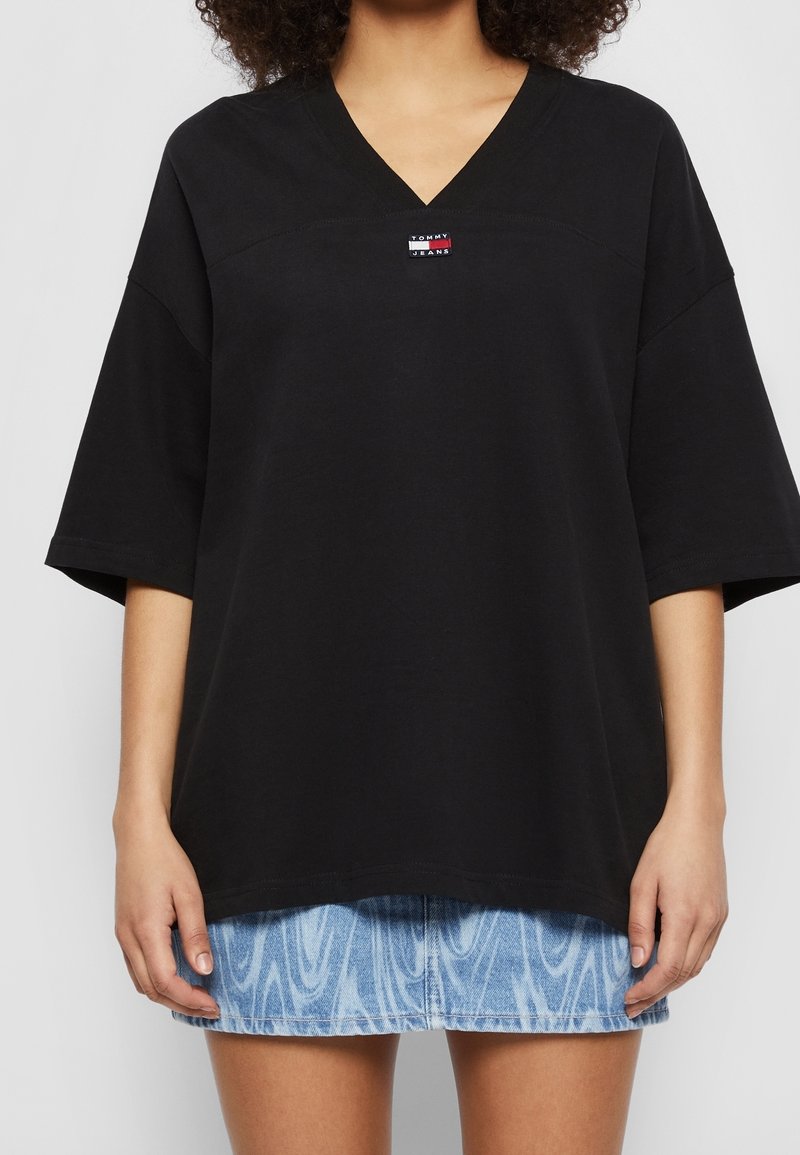 T-shirt oversized nera a scollo a V in cotone con un piccolo dettaglio logo rosso, bianco e blu sul petto; indossata con una gonna di jeans blu chiaro.