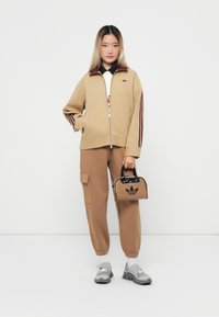 Giacca beige con zip e strisce bordeaux, t-shirt bianca, pantaloni jogging marroni con tasche e borsa nera con logo Adidas. Sneakers grigie.