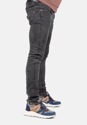 Jambes portant un jean noir ajusté et des baskets grises, bleues et marron avec des semelles blanches, se tenant sur un fond clair uni.