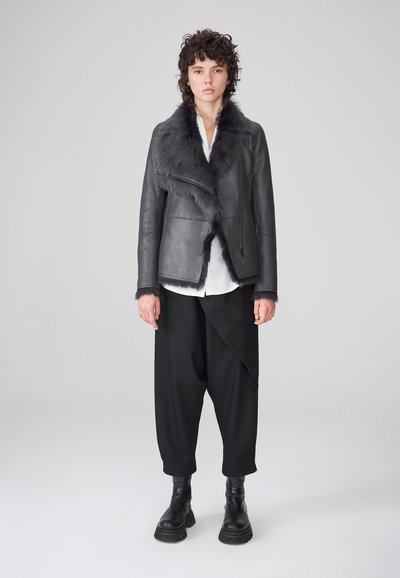 Chaqueta de cuero gris con un cuello de piel sintética, diseño asimétrico y cierre de cremallera, combinada con pantalones negros holgados y botas negras.