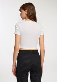 Biały dopasowany crop top z krótkimi rękawami, wykonany z elastycznego materiału. Noszony z czarnymi spodniami w prążek o wysokim stanie. Pokazany widok z tyłu.