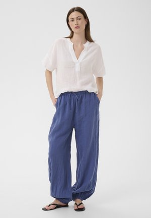 Frau steht mit den Händen in den Taschen, trägt eine lockere weiße Bluse, weite blaue Hosen mit Kordelzug und schwarze Flip-Flops auf weißem Hintergrund.