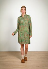 Woman wearing a green paisley-patterned long-sleeve shirt dress, beige socks, and brown slippers standing on wooden floor.